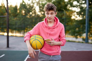 portrait of caucasian basketballer boy with smartphone and ball in hands, have reat after game