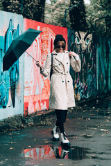 young stylish woman wearing long beige coat, white boots, black hat, umbrella and backpack posing through the city streets. Selective focus, grain