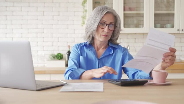 Serious worried old senior woman reading paper bill pay online at home, managing bank finances, calculating taxes, fees, high rental rates, planning loan debt pension payment sit at kitchen table.
