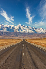 Portrait size shot of a straight empty highway leading to the snowy peaks of The Kuray mountain range. Beautiful blue cloudy sky as a background. Altai mountains, Siberia, Russia