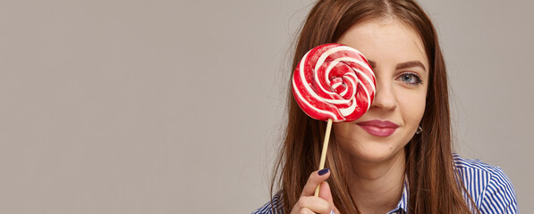 happy modern teenage woman smiling and hiding behind a lollipop candy over grey background.