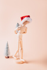 Wooden mannequin in red Santa hat is carrying tiny christmas tree, concept of preparation for Xmas holidays, going home for festive time concept