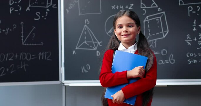 Portrait of cute Caucasian small teen clever girl with copybook standing in classroom at blackboard with math formulas and smiling to camera. Smart pretty little schoolgirl smile. Mathematics.