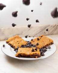 Blondies on white plate with chocolate chips falling on top. Decorated with milk, chocolate chips and Nutella. Delicious pastries out of the oven and ready to eat. Perfect for a bakery or online store