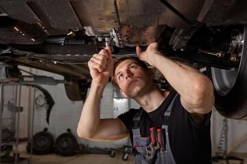 Obraz na płótnie Canvas young male car mechanic in uniform checking car in automobile service with lifted vehicle, handsome hardworking guy working under car condition on lifter. automotive car repair concept