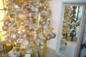 A Christmas tree with a Golden decoration is reflected in the mirror. New year, home decoration. Large window with yellow warm light, comfort in the living room, gift boxes