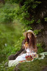 Cute red girl in straw hat on a flowering clearing at a picnic with fruit