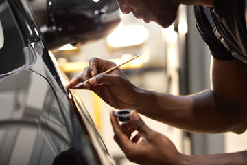 young afro auto service worker is painting car details, use small brush and paint, apply it on surface of black automobile