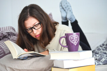 Young woman with short brown hair and glasses in cozy clothes reading a book in bed at home. Pile...