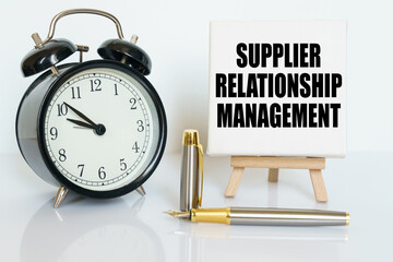 On the table there is a clock, a pen and a stand with a card on which the text is written - SUPPLIER RELATIONSHIP MANAGEMENT