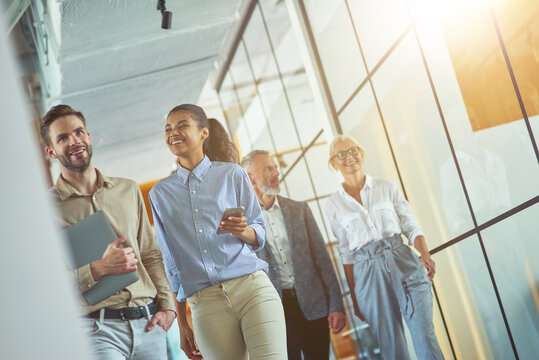 Two Young Happy Diverse Male And Female Coworkers Discussing Something And Smiling While Walking With Colleagues In The Modern Office Or Coworking Space