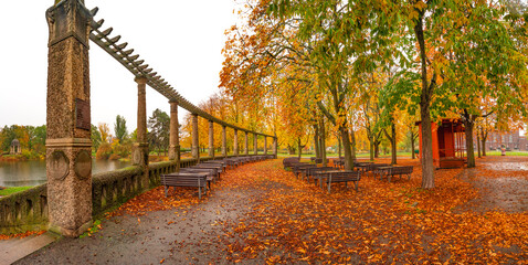 Panorama over lonely empty benches under old chestnut trees and old columns with fallen red and orange leaves in the city park in Autumn colors in rainy day, Magdeburg, Germany.
