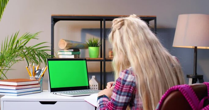 Back view on Caucasian teen girl sitting at table studying online at laptop computer. Schoolgirl writing in copybook and doing homework. Lockdown learning. Green screen. Chroma key. Quarantine. Rear.