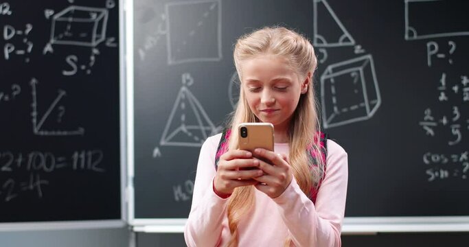 Cheerful pretty Caucasian schoolgirl standing in school at blackboard with math and chatting on smartphone Teenage beautiful schoolgirl tapping and texting message on mobile phone at mathematics class