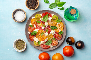 Italian caprese salad with varicolored tomatoes, mozzarella cheese, basil leaves, spices and olive oil in plate on concrete background