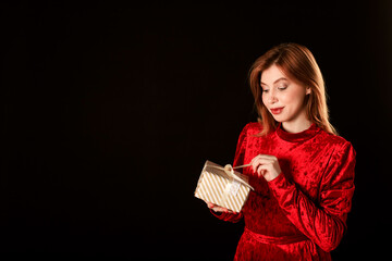 A girl in a red dress on a black background holds a gift in her hand and opens it.