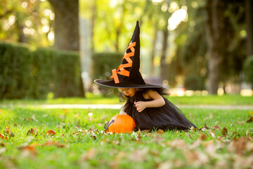 A cute little girl in a witch costume walks in the street with a candy in the shape of a pumpkin. The concept of Halloween