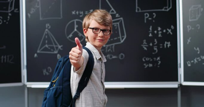 Portrait of cute happy Caucasian boy with backpack standing in school at blackboard with math formulas and giving thumb up. Teenage schoolboy in glasses smiling at mathematics class.