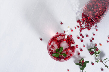 Cold cocktail with lingonberry, ice cubes, straws, scattered berry in transparent glass on soft light white wood table with copy space, top view.
