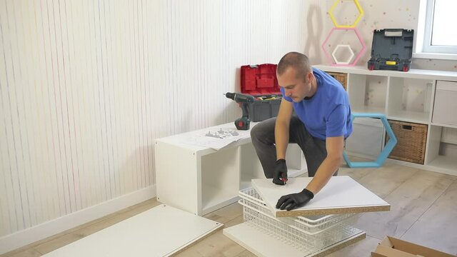 A guy in a blue t-shirt assembles white furniture in a room