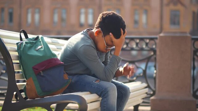Side view of sad mixed-race schoolboy sitting alone on bench outdoors