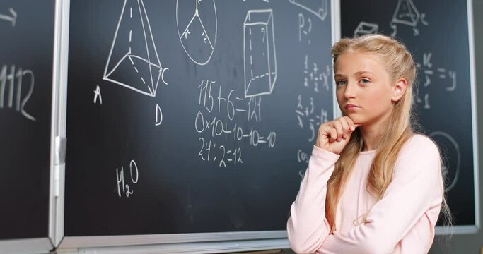 Caucasian small teen girl standing at blackboard in classroom, thinking and deciding math formulas and equations. Teenage smart schoolgirl studying mathematics at chalkboard. Algebra or geometry class