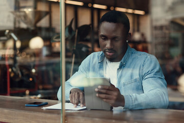 Purposeful black male entrepreneur uses tablet pc while sitting in cafe with coffee near window.