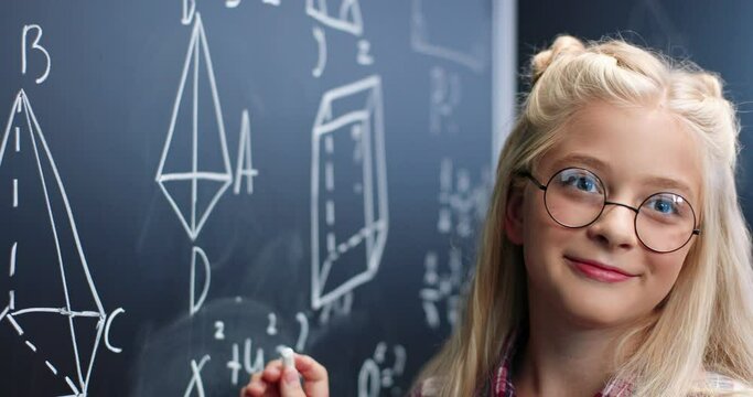 Close up of Caucasian small teen girl in glasses standing at blackboard in classroom and deciding math formulas and equation. Teenager schoolgirl studying mathematics at chalkboard. Geometry. Portrait