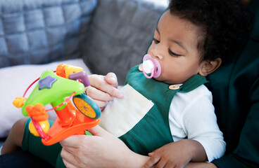 African American baby boy with pacifier is interesting his favorite toys while sit on mother's lap in living room. Portrait of a cute little happy black baby boy with mother at home, Family concept.