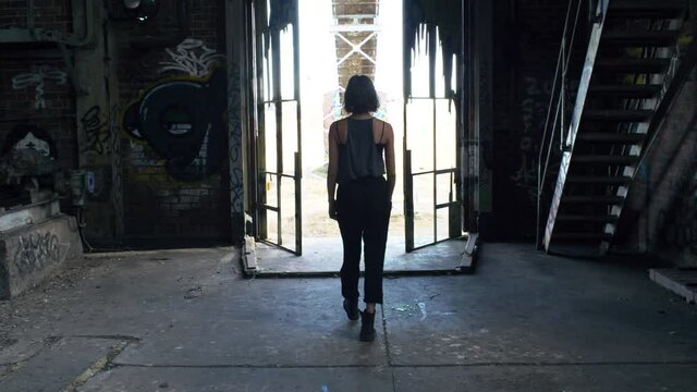 Woman In Grunge Fashion Clothing Walking Towards Exit Of A Rundown Warehouse - medium shot