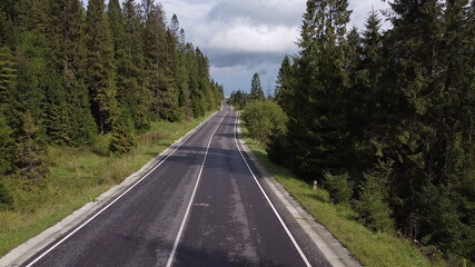 Naklejka premium Aerial low air flight over mountain perfect asphalt highway road and meadow at sunny summer morning. Near green trees. Fast horizontal movement with sun rays.
