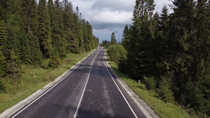 Aerial low air flight over mountain perfect asphalt highway road and meadow at sunny summer morning. Near green trees. Fast horizontal movement with sun rays.