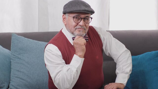 adulto mayor bien parecido con sombrero y anteojos afirmando sentado en una habitaci&oacute;n de casa feliz y tranquilo
