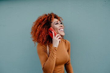 Cheerful young woman with afro hair talking over mobile phone while standing against wall