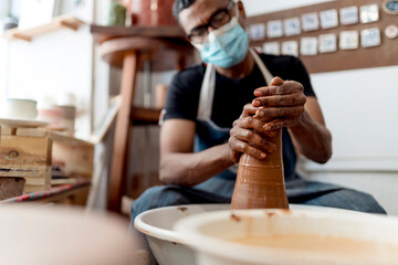Fototapeta na wymiar Male potter wearing mask molding shape to clay while sitting in workshop