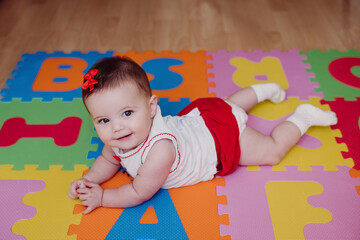 Cute smiling baby girl lying on colorful puzzle playmat at home