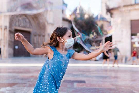 Carefree girl wearing mask running on street in city playing with soap bubble