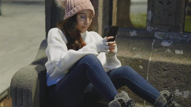 niña bonita,alegre vestida con ropa de invierno sentada tomándose fotos y mandando mensajes en el celular al aire libre