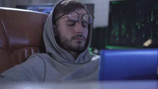 Close-up portrait of Caucasian young man waking up, putting on eyeglasses and start typing on laptop keyboard. Serious exhausted programmer coding online or hacker hacking web site.