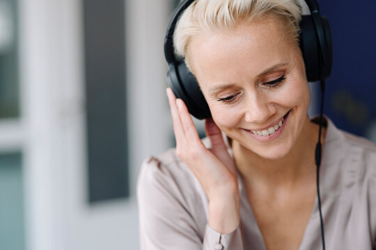 Close-up of smiling businesswoman listening music over headphones looking down in office