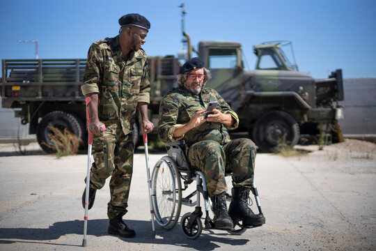 Injured army soldier holding crutches while standing with disabled colleague on wheelchair looking at smart phone during sunny day