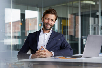 Smiling businessman with hands clasped sitting by desk in office