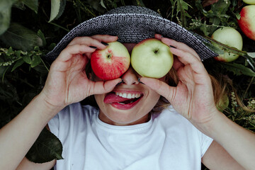 Close-up of cheerful woman holding apples in front of eyes while lying at in orchard