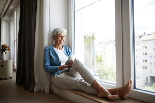 Happy senior female using digital tablet while sitting on window at home