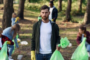 Good-looking bearded man shows a full rubbish pack in background of his friends volunteers picking...