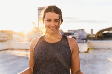 Smiling female athlete standing on building terrace against sky during sunset
