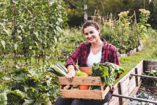Smiling young woman with vegetables in crate sitting against plants at garden
