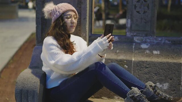 ni&ntilde;a bonita,alegre vestida con ropa de invierno sentada tom&aacute;ndose fotos y mandando mensajes en el celular al aire libre