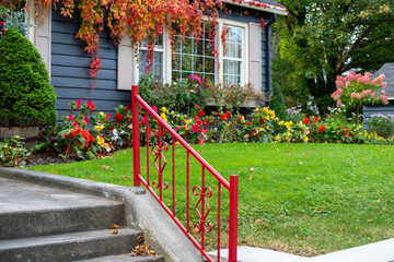 Fototapeta na wymiar A bright red metal handrail leading up four concrete steps to a walkway. There's a green lawn, red and yellow flowers, red vines and large green shrub. A group of tall green trees in the background.