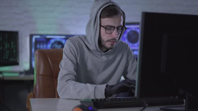 Purposeful man in eyeglasses putting on black gloves and typing on computer keyboard. Portrait of male Caucasian hacker stealing information from office or hacking security password.
